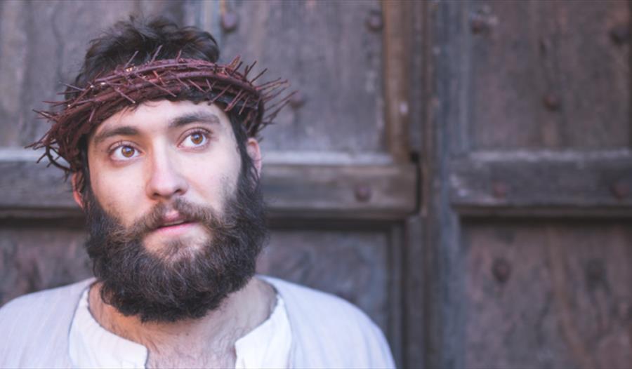 A bearded man wearing white with a crown of thorns on his head plays Jesus in the Chester City Passion. He is looking foward, and behind him is an old wooden door.